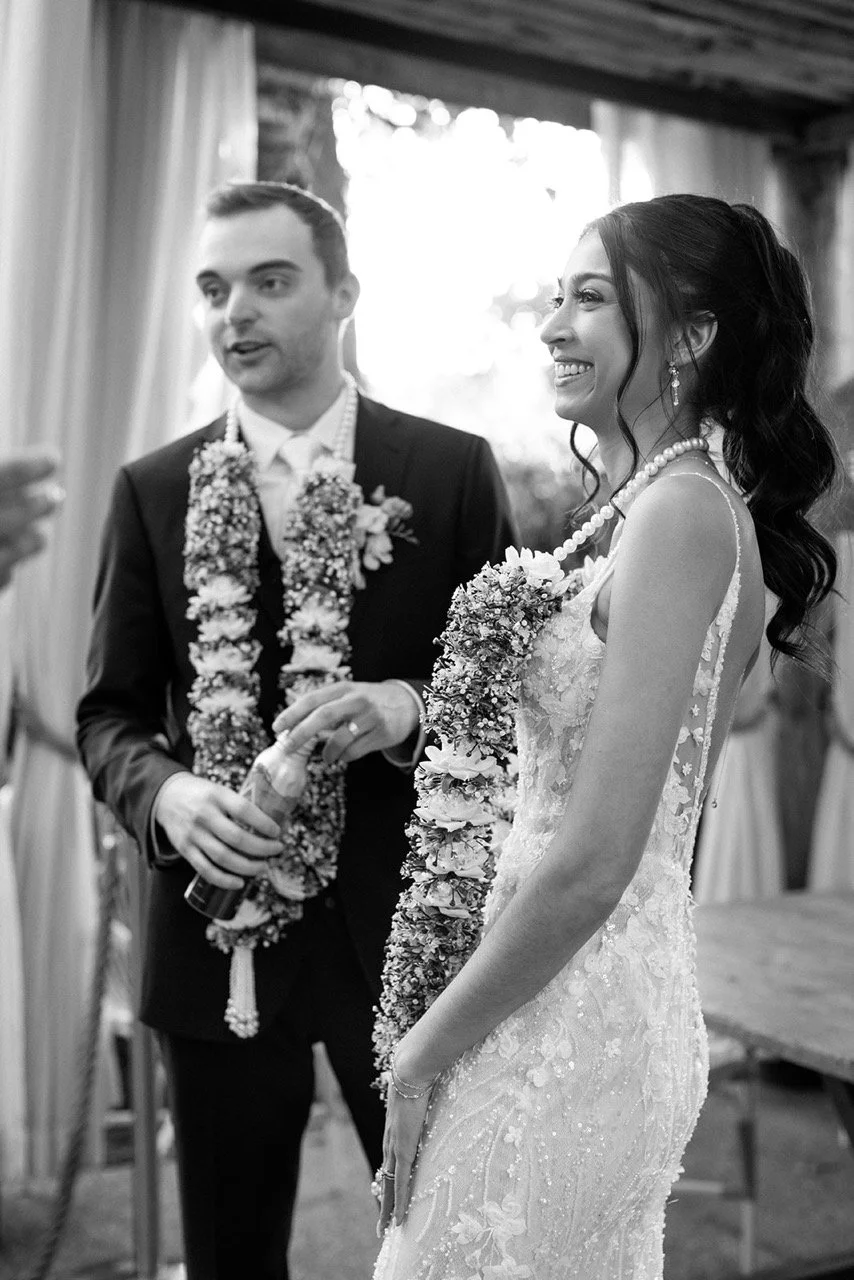 Black and white photo of a bride and groom at their wedding ceremony, wearing floral leis. The bride is smiling with her hair in loose curls, wearing a lace dress and pearl necklace. The groom is speaking, dressed in a suit with a flower boutonniere