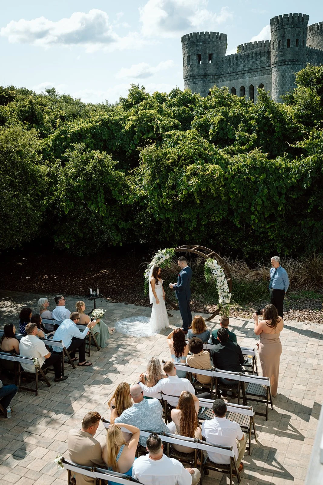 An outdoor wedding ceremony taking place in front of a lush green hedge and a castle-like structure with towers. The bride and groom stand under a floral arch, exchanging vows as guests sit on benches and chairs watching.