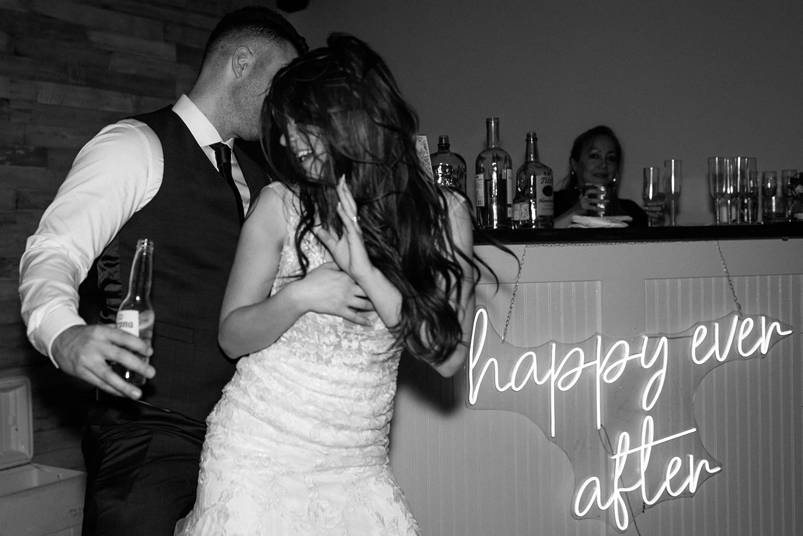 A couple dancing at a wedding reception, with a woman in the background holding a drink behind a bar adorned with a neon sign that reads "happy ever after".