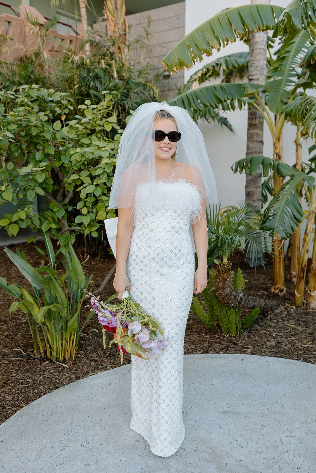 Woman in a white wedding dress, veil, and sunglasses holding a bouquet of flowers outdoors