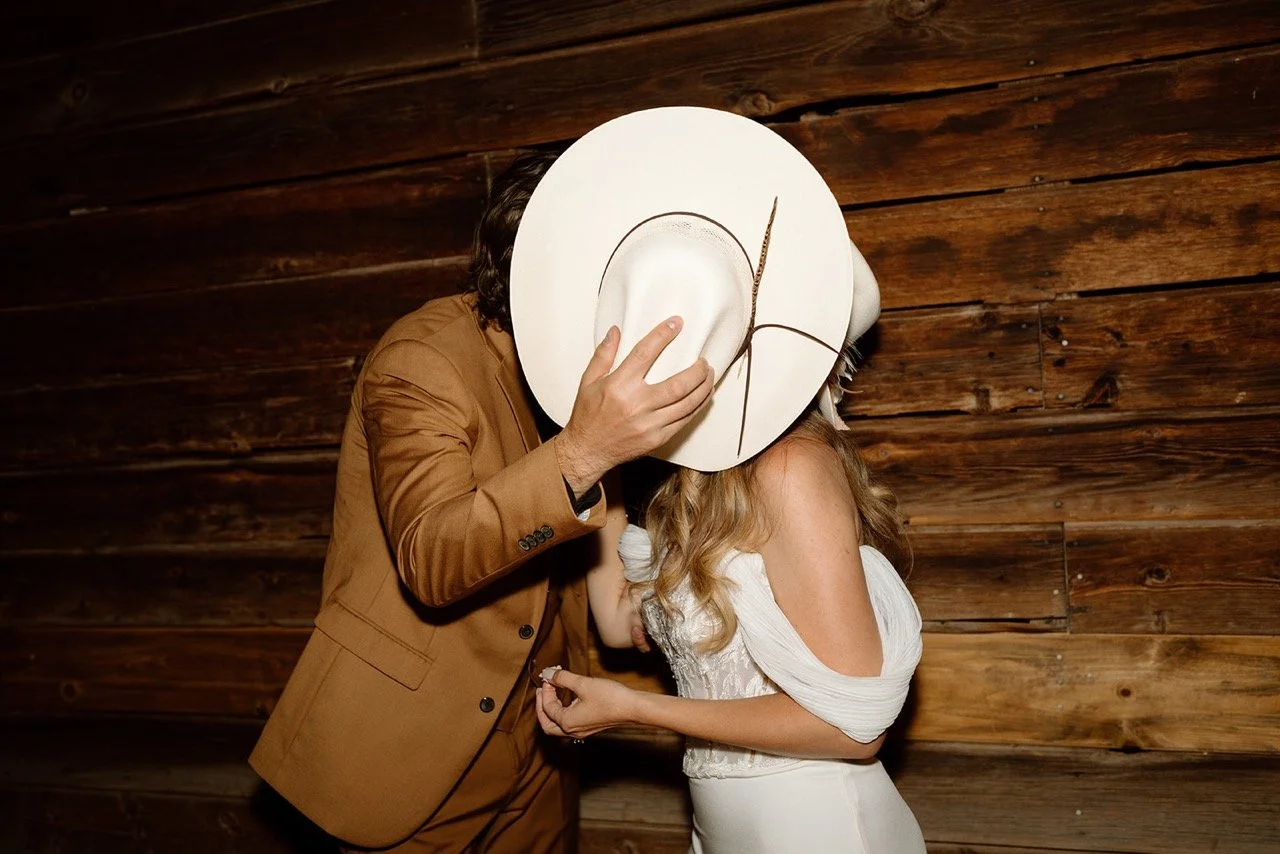 Western Wedding at Saguaro Lake Guest Ranch - bride and groom kissing behind cowboy hat next to rustic barn