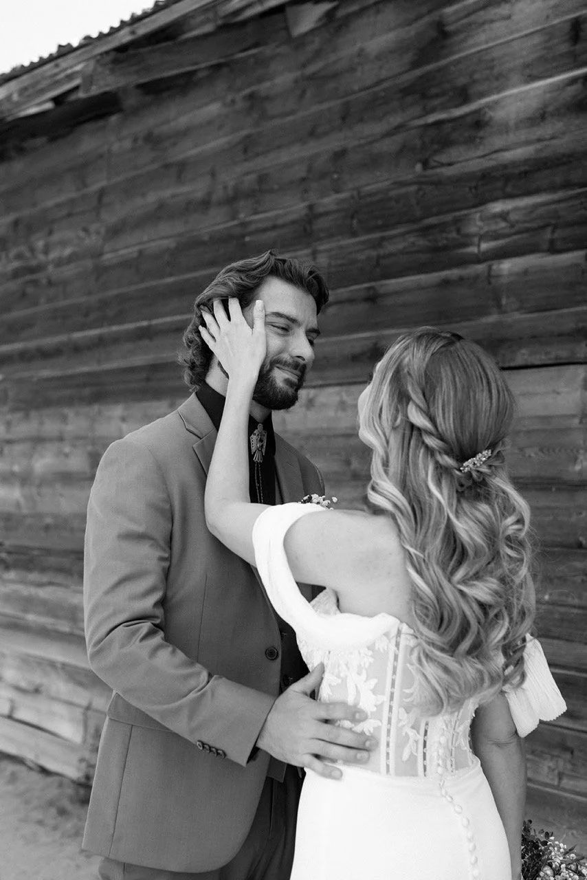 A black and white photo of a couple on their wedding day. The woman, with long, wavy hair, is touching the man's face and the man, dressed in a suit, is smiling with his eyes closed. They are standing close to a wooden wall.