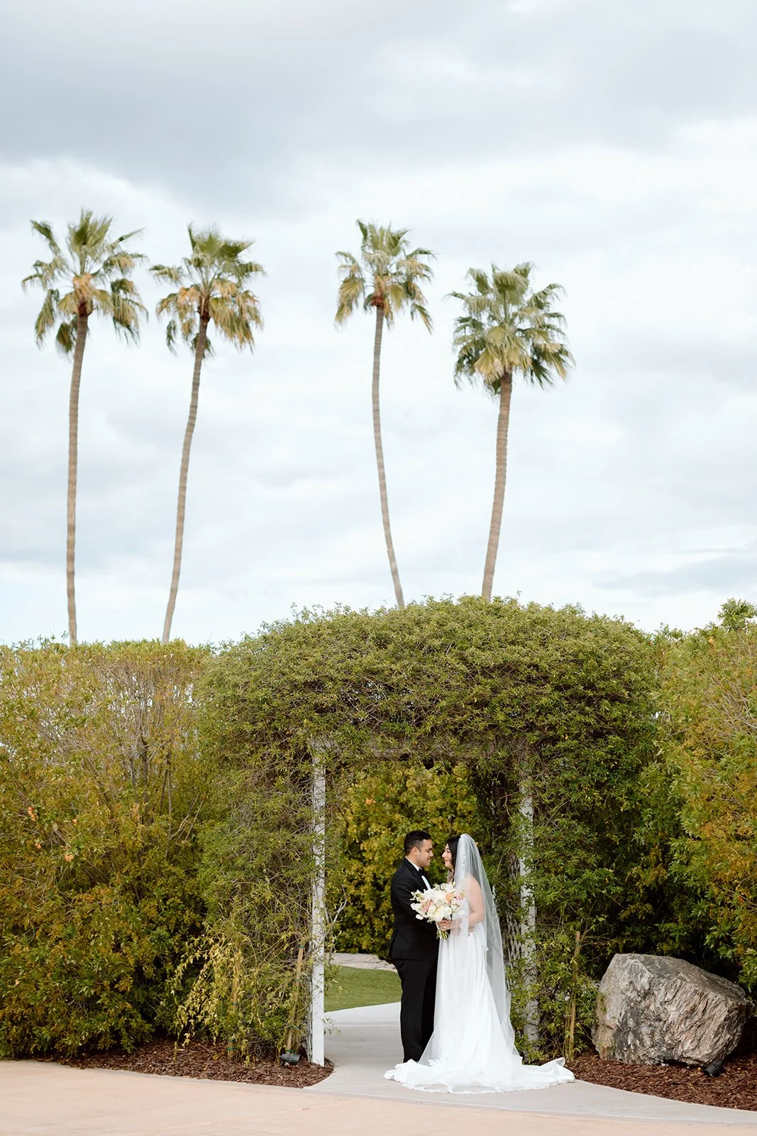 A bride and groom standing under a lush green archway, holding a bouquet, with tall palm trees and an overcast sky in the background.