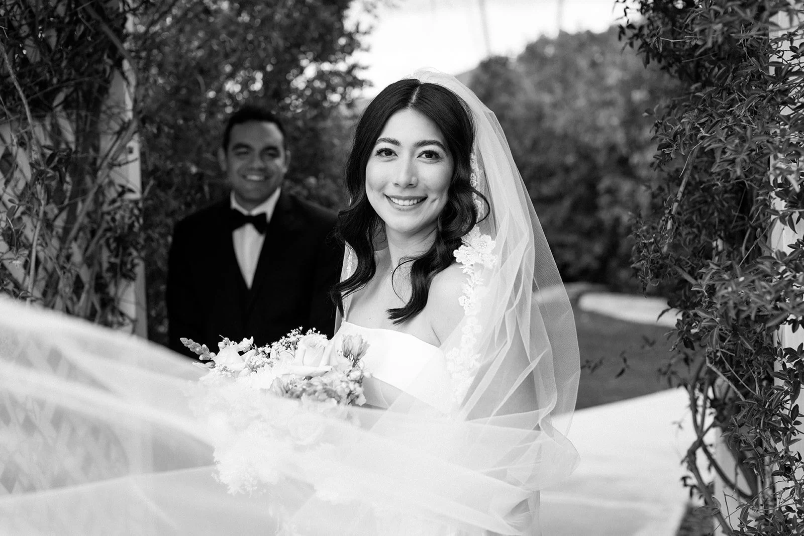 Black and white wedding photo of a smiling woman in a bridal gown holding a bouquet, with a man in a tuxedo smiling behind her, surrounded by greenery.