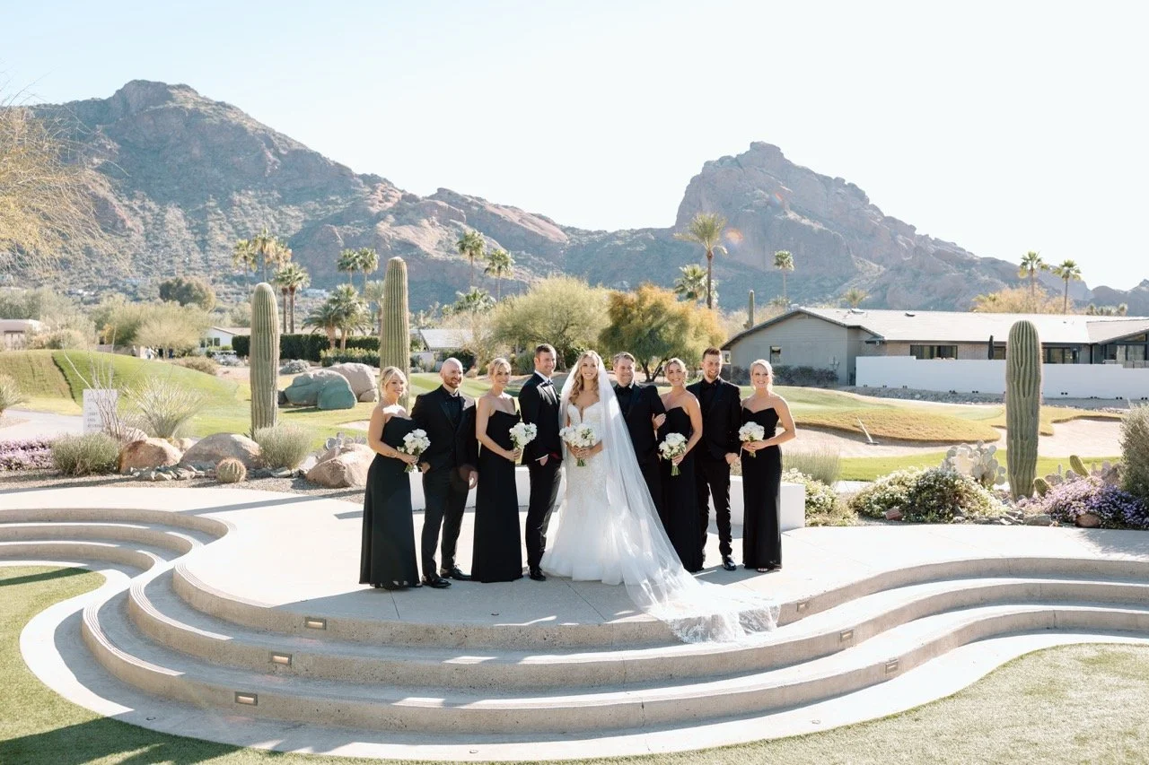 Group of wedding party members in formal attire, including the bride in a white wedding dress and veil, and bridesmaids and groomsmen in black dresses and suits, standing on a tiered outdoor stage with mountains and desert landscape in the background