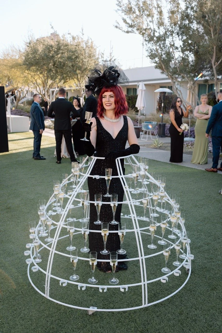 A woman dressed in a black evening gown with long black gloves stands inside a white metal balloon holder structure at an outdoor event. She has red hair, is smiling, and holds a glass of champagne. The background shows other guests in formal attire,