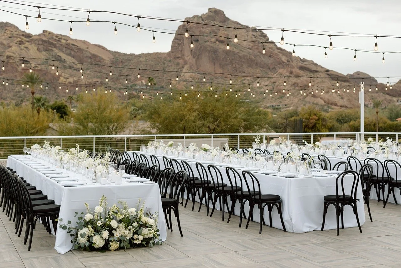 Outdoor event space with long white-draped banquet tables, black chairs, floral centerpieces, string lights, and mountain scenery in the background.