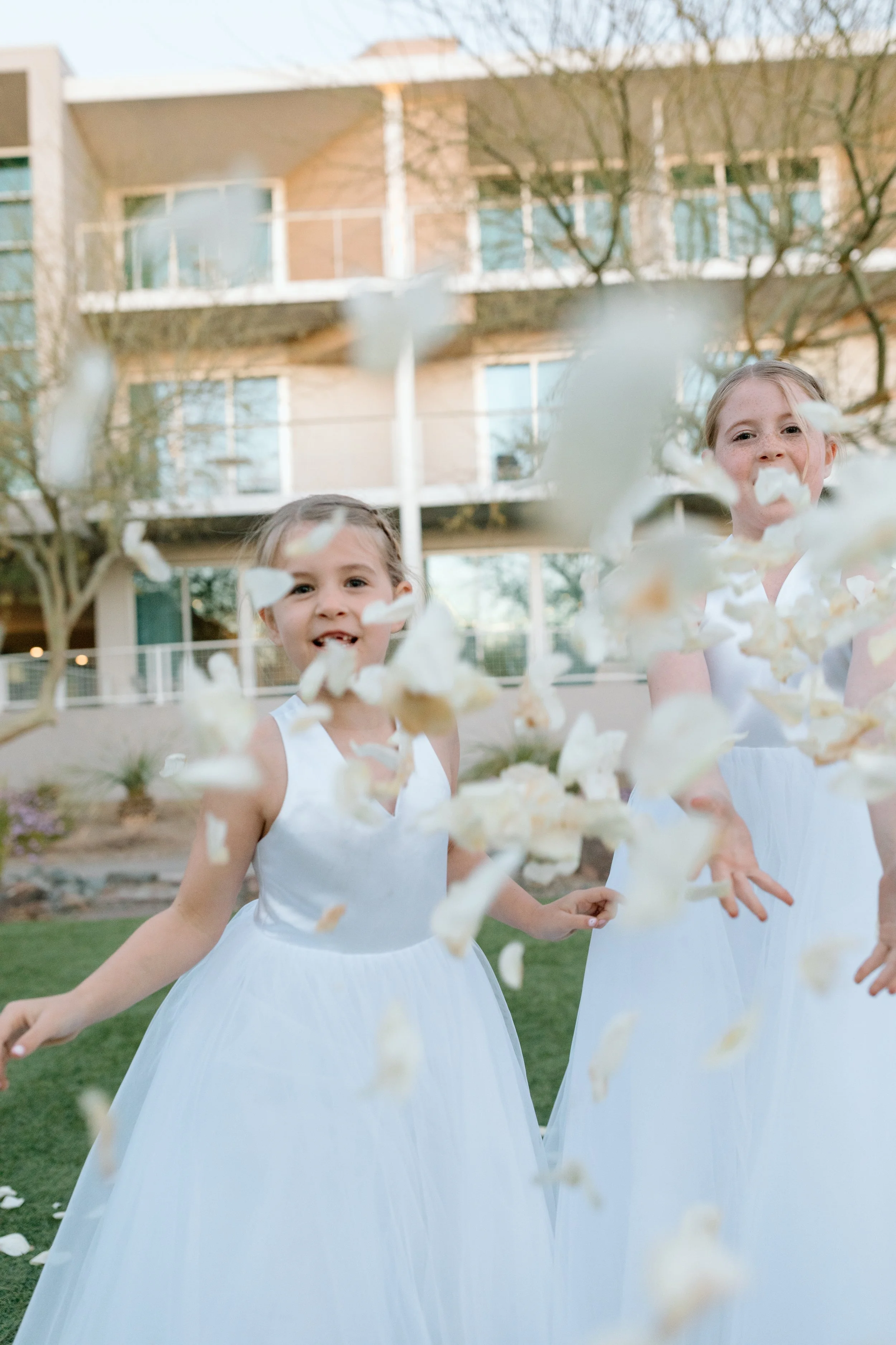 Two young girls in white dresses playing with flower petals outdoors in front of a modern apartment building.