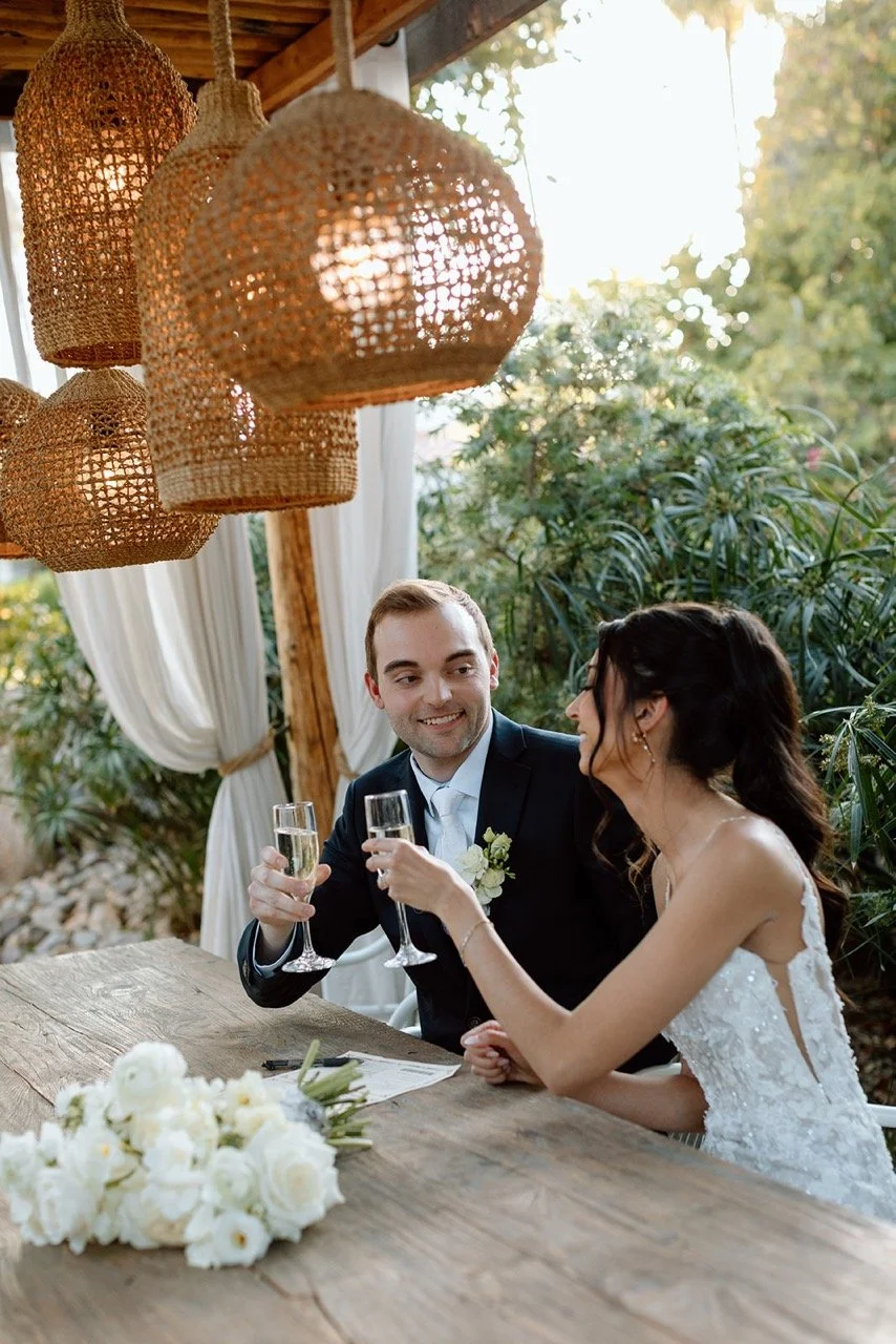 A bride and groom sitting at a rustic wooden table during their wedding celebration, clinking champagne glasses, with wedding flowers on the table and wicker pendant lights hanging above.