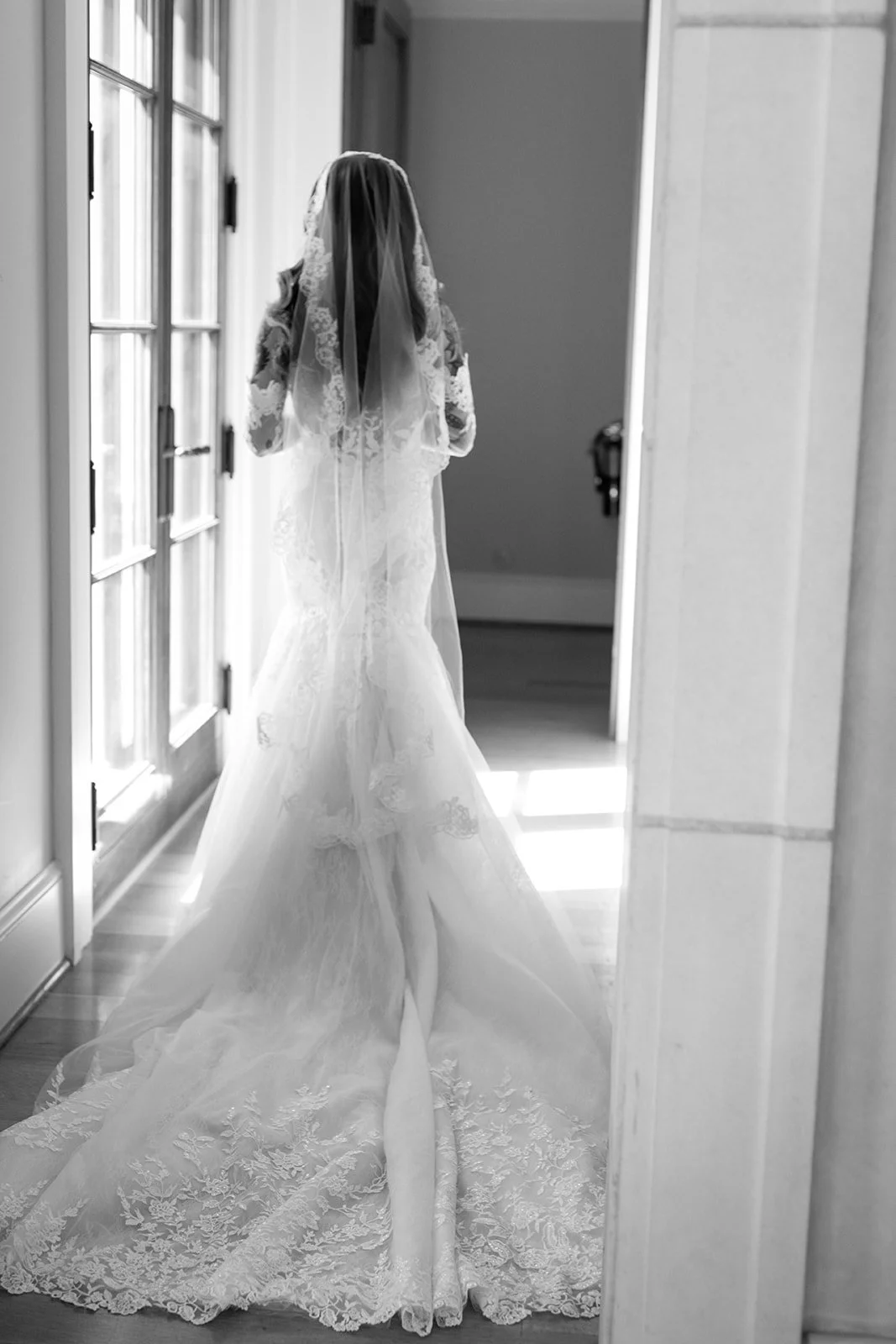 A woman in a wedding dress and veil standing in front of a window, seen from behind in black and white.