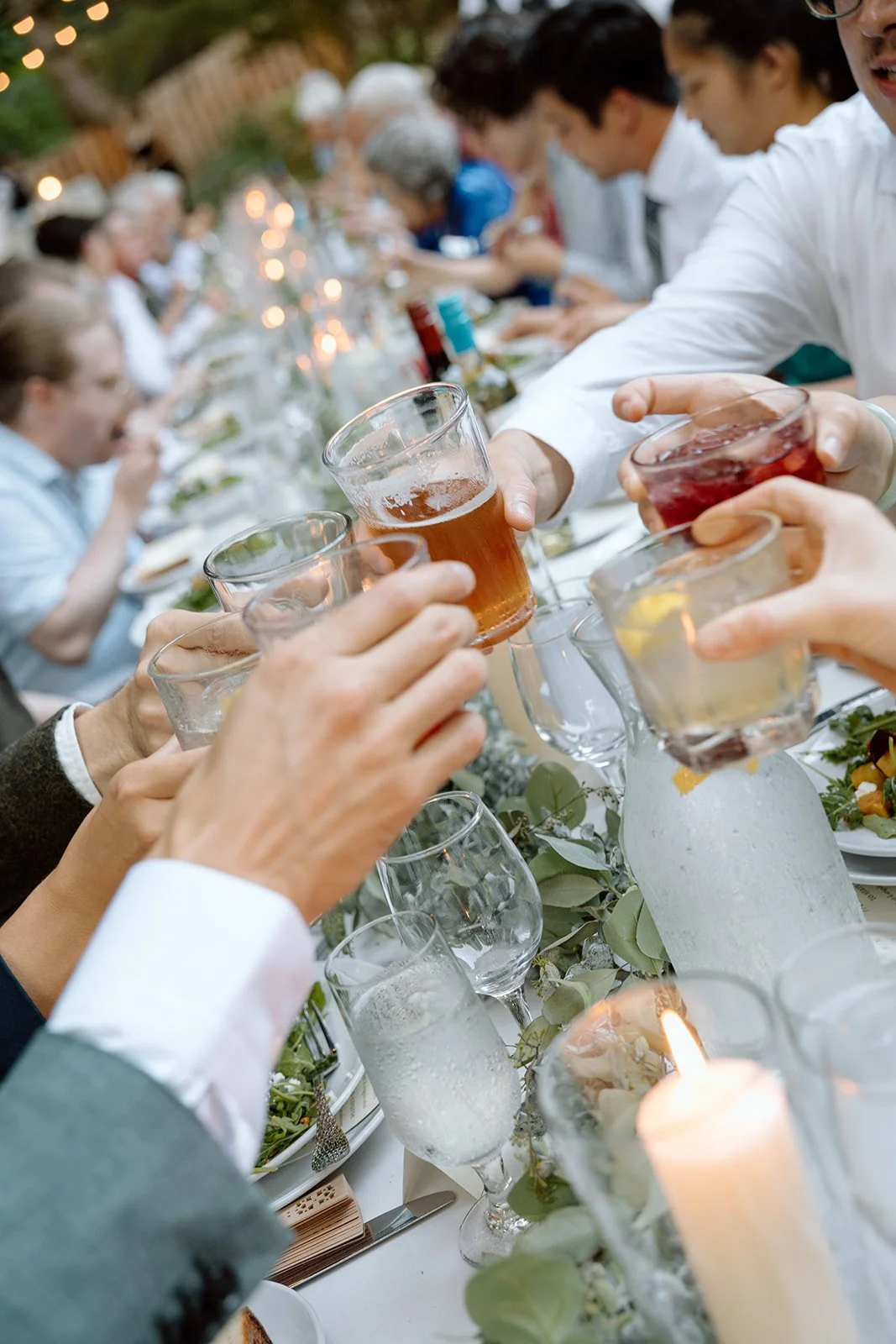 People raising glasses for a toast at a festive dinner gathering, with a long table set with plates, glasses, candles, and greenery.