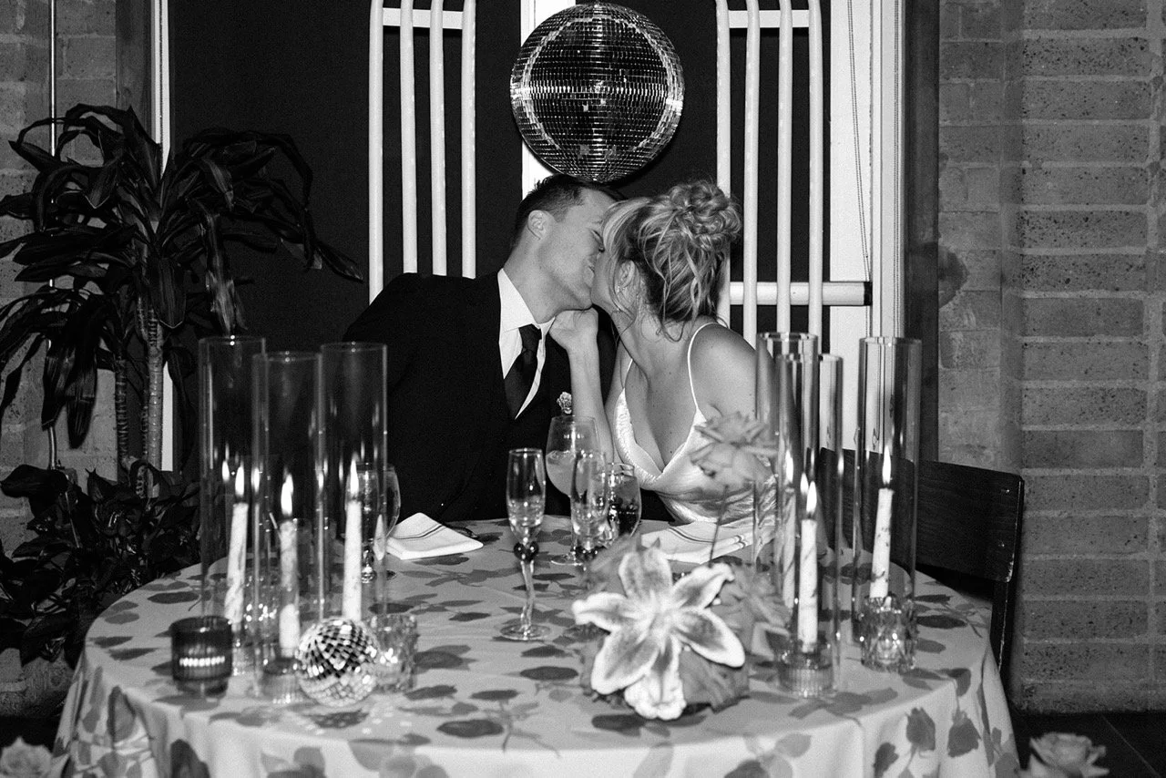 A black and white photo of a couple sharing a kiss at a decorated table with flowers, candles, and glasses, in a dimly lit indoor setting.