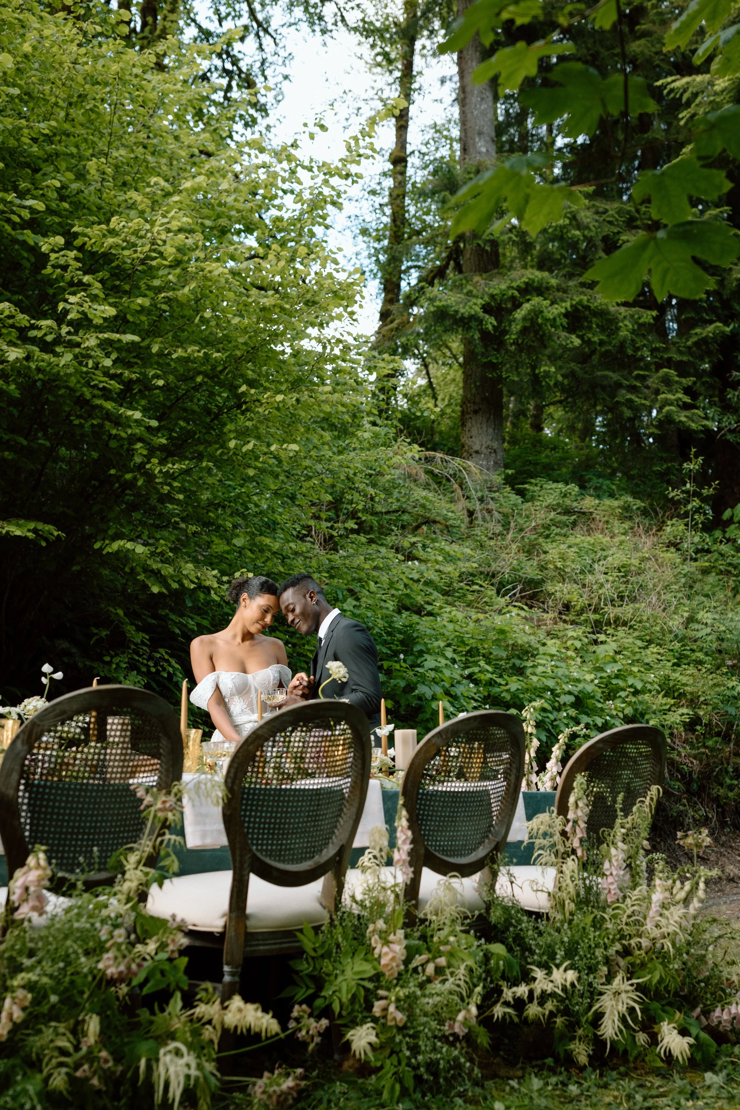 A bride and groom standing close together at an outdoor wedding reception table surrounded by lush green trees and foliage.