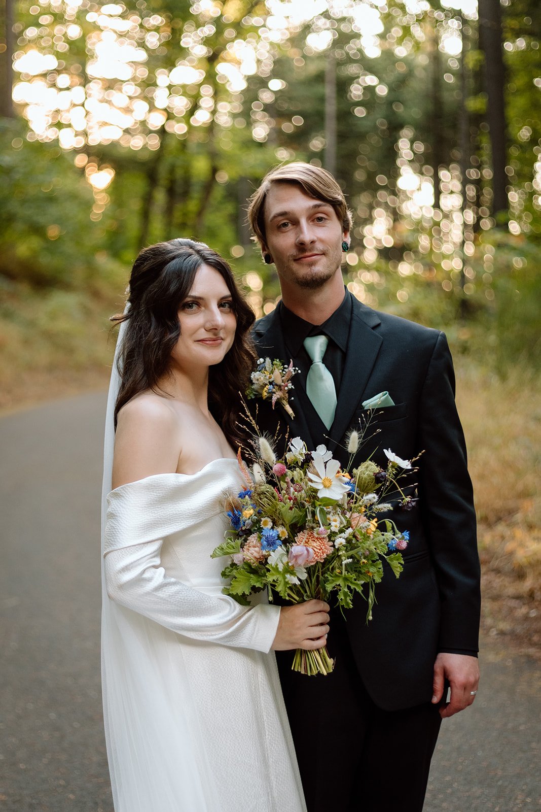 A bride and groom standing together outdoors in a wooded area, with the bride holding a colorful bouquet of flowers, both dressed in wedding attire, looking at the camera.