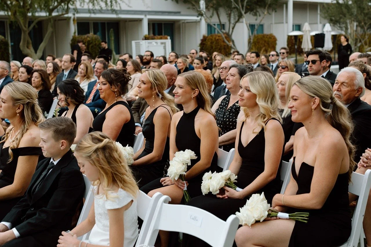Attendees sitting in rows at an outdoor wedding reception, many women holding white bouquets, dressed in formal black and white attire.
