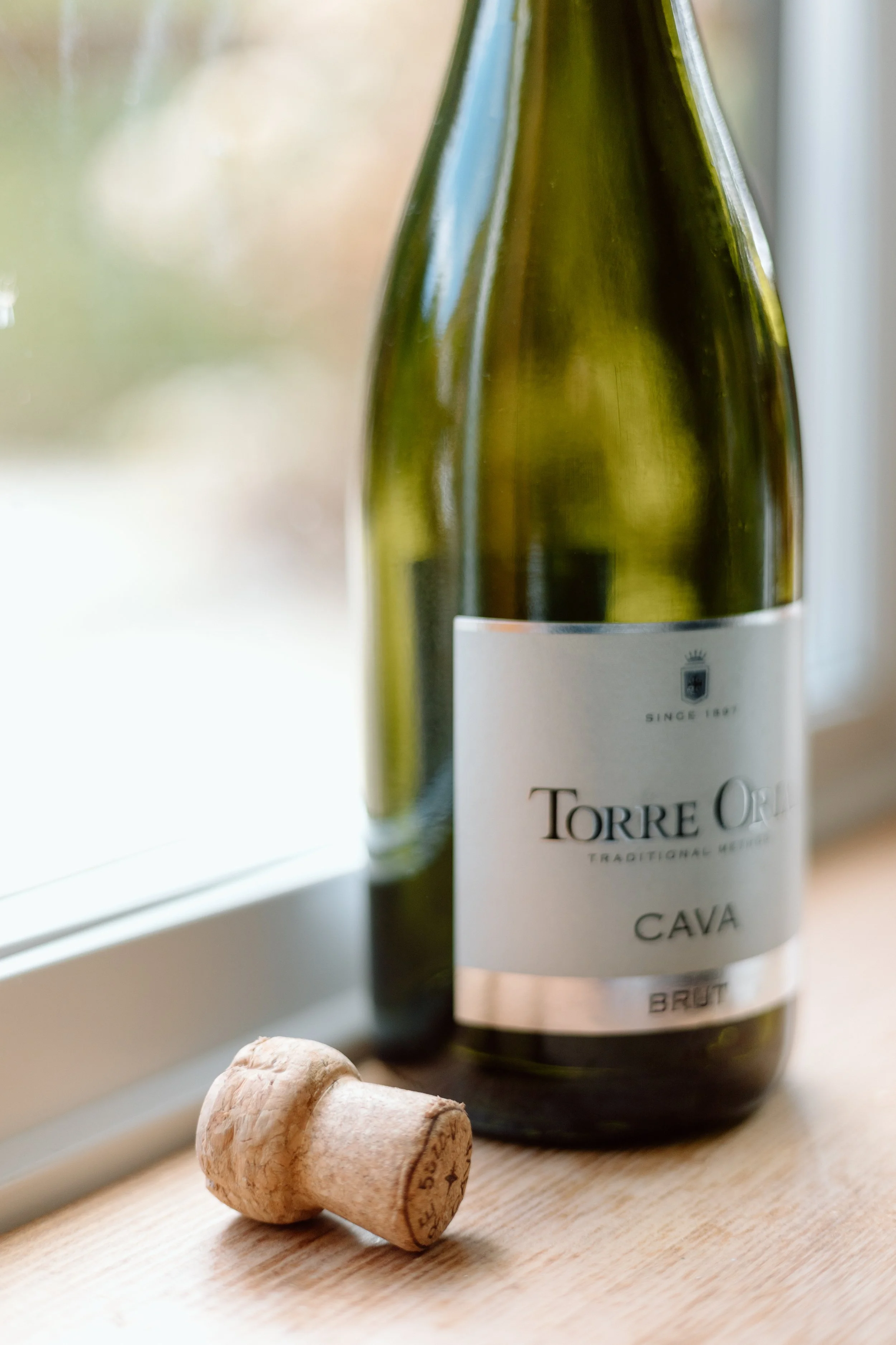 A bottle of Torre Ori Cava Brut sparkling wine and a cork lying on a wooden surface near a window.