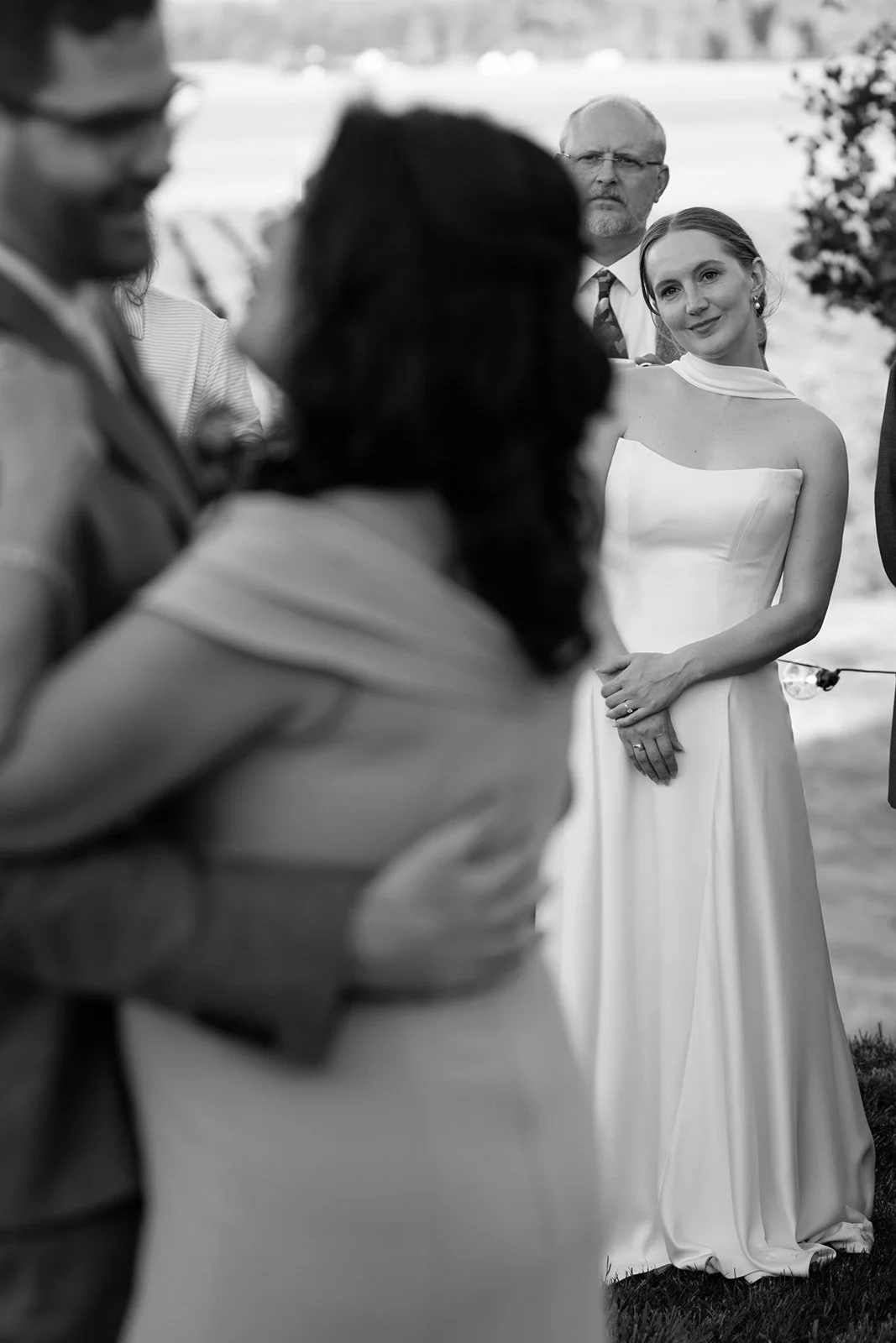A black and white photo of a wedding ceremony outdoors, featuring a woman in a strapless white wedding gown, a man with glasses, and other guests in the background.