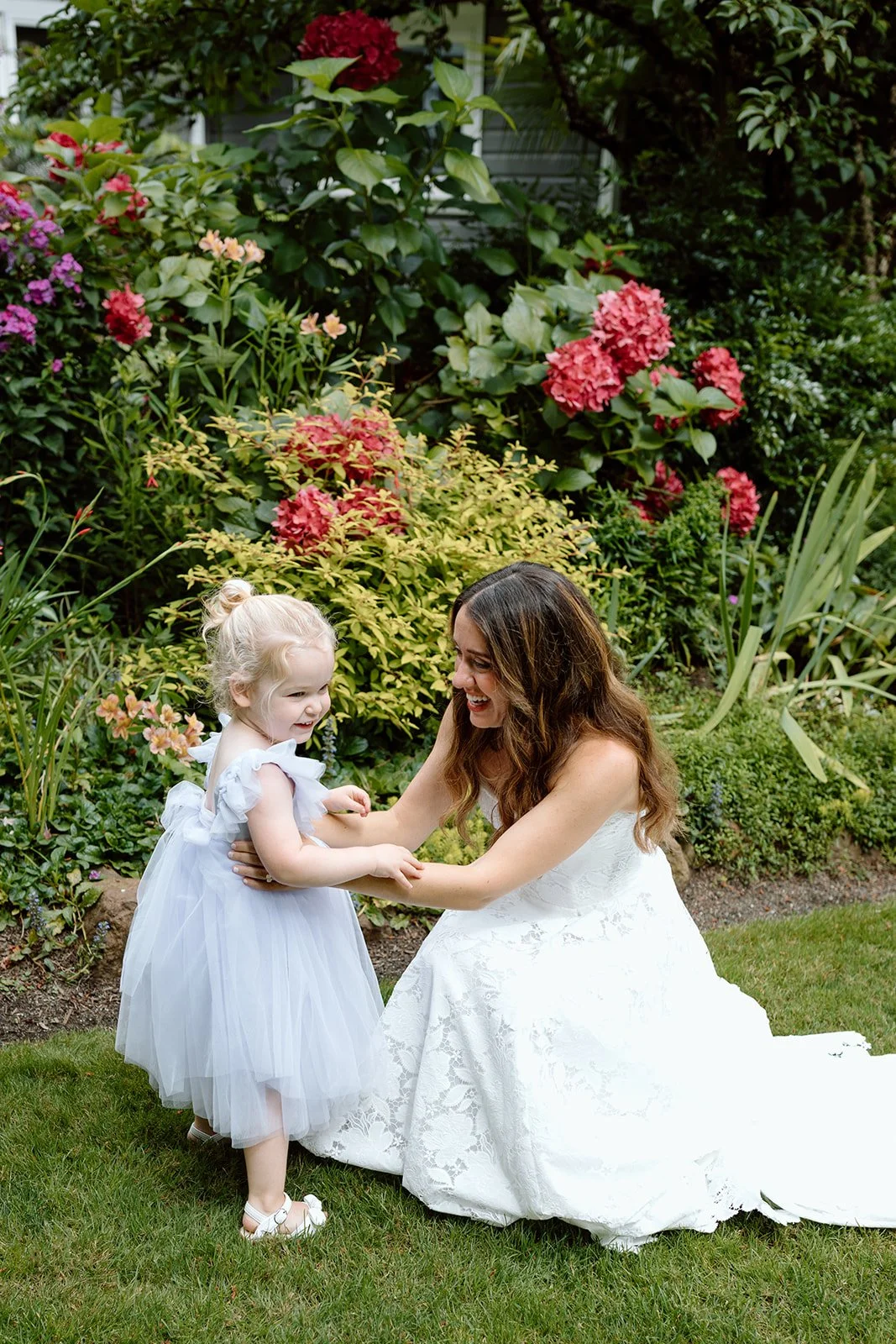 A woman in a white lace wedding dress and a young girl in a white dress with a tulle skirt smiling and playing together in a garden with colorful flowers and greenery.