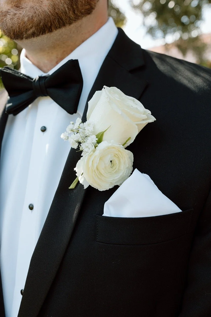 Close-up of a man in a black tuxedo with a white dress shirt, black bow tie, and white boutonniere consisting of a white rose and small white flowers on the left lapel.