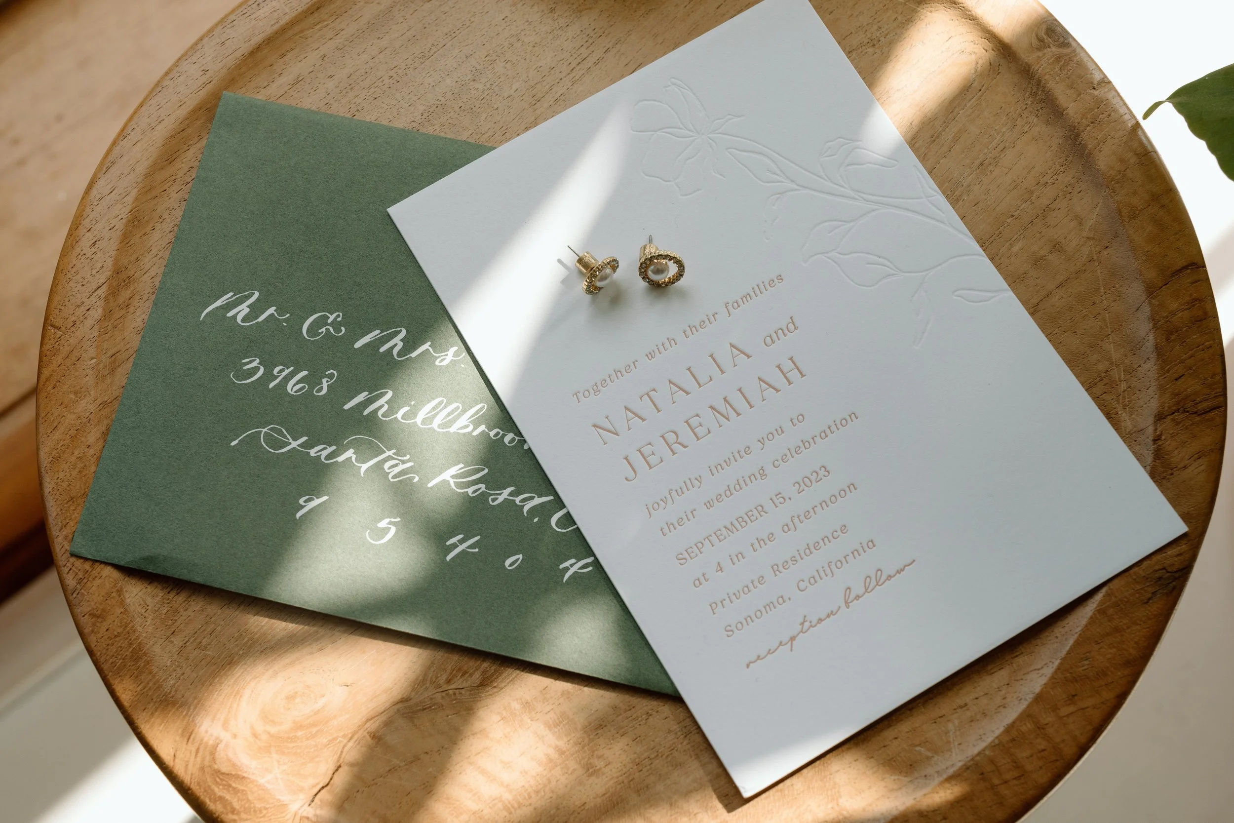 Wedding invitation card with Eucalyptus design, gold text, and a pair of pearl earrings on a wooden table.