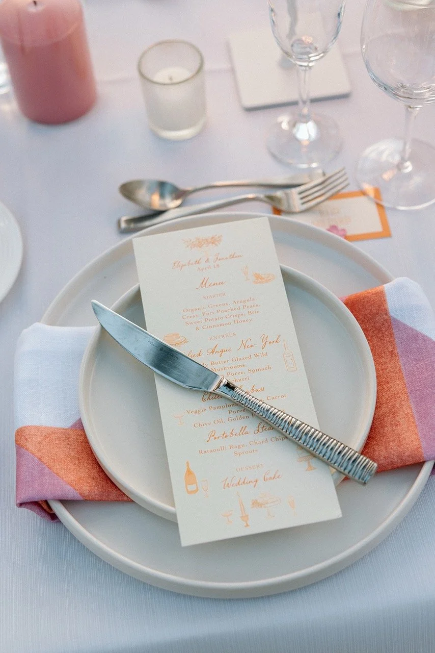 Elegant table setting with a white plate, a menu card, a knife, a pink and white napkin, and glassware on a white tablecloth