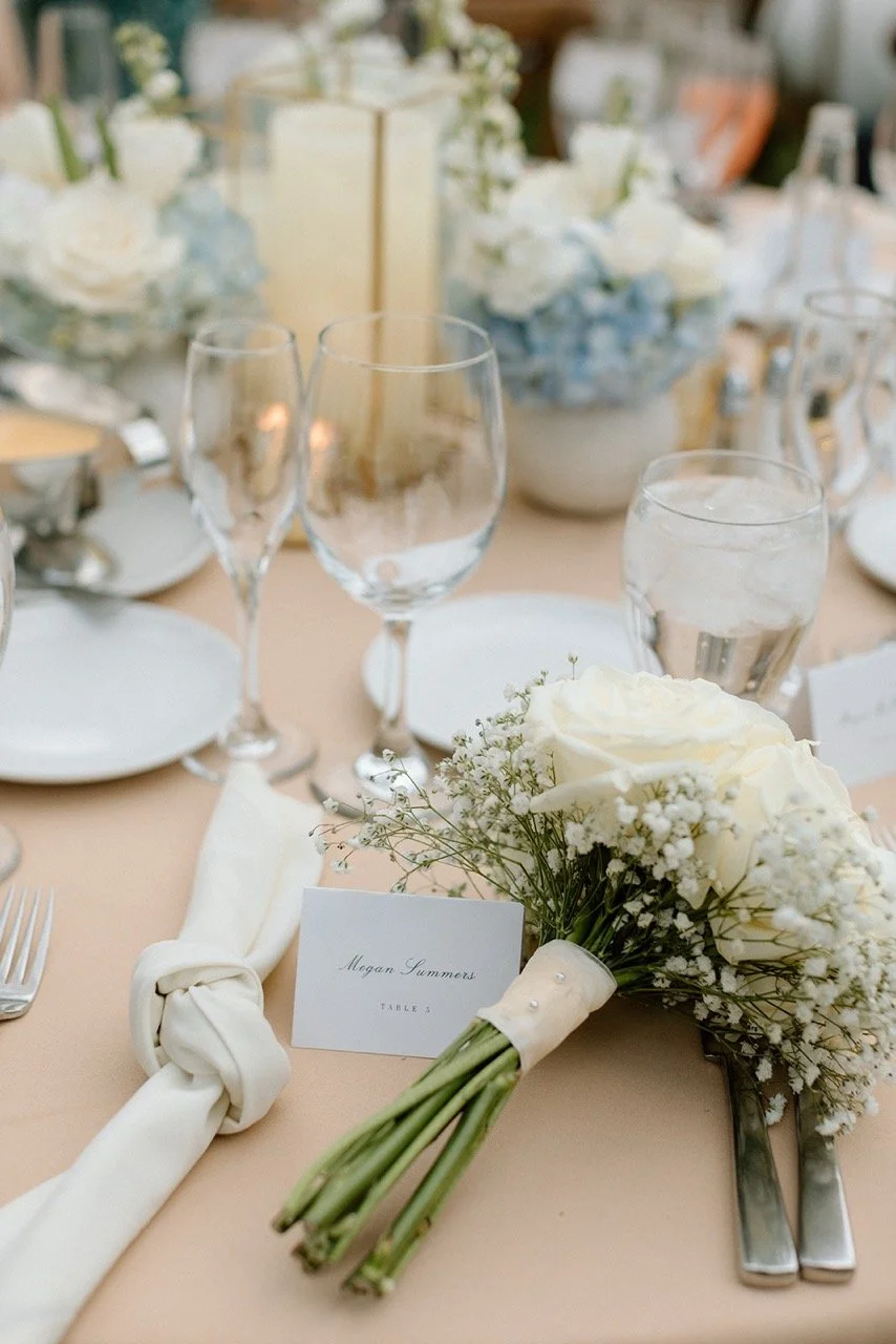 Wedding table setting with a bouquet of white roses and baby's breath, a name card reading 'Megan Summers, Table 3', white napkin tied with a knot, and glassware including wine glasses and water glasses on a beige tablecloth.