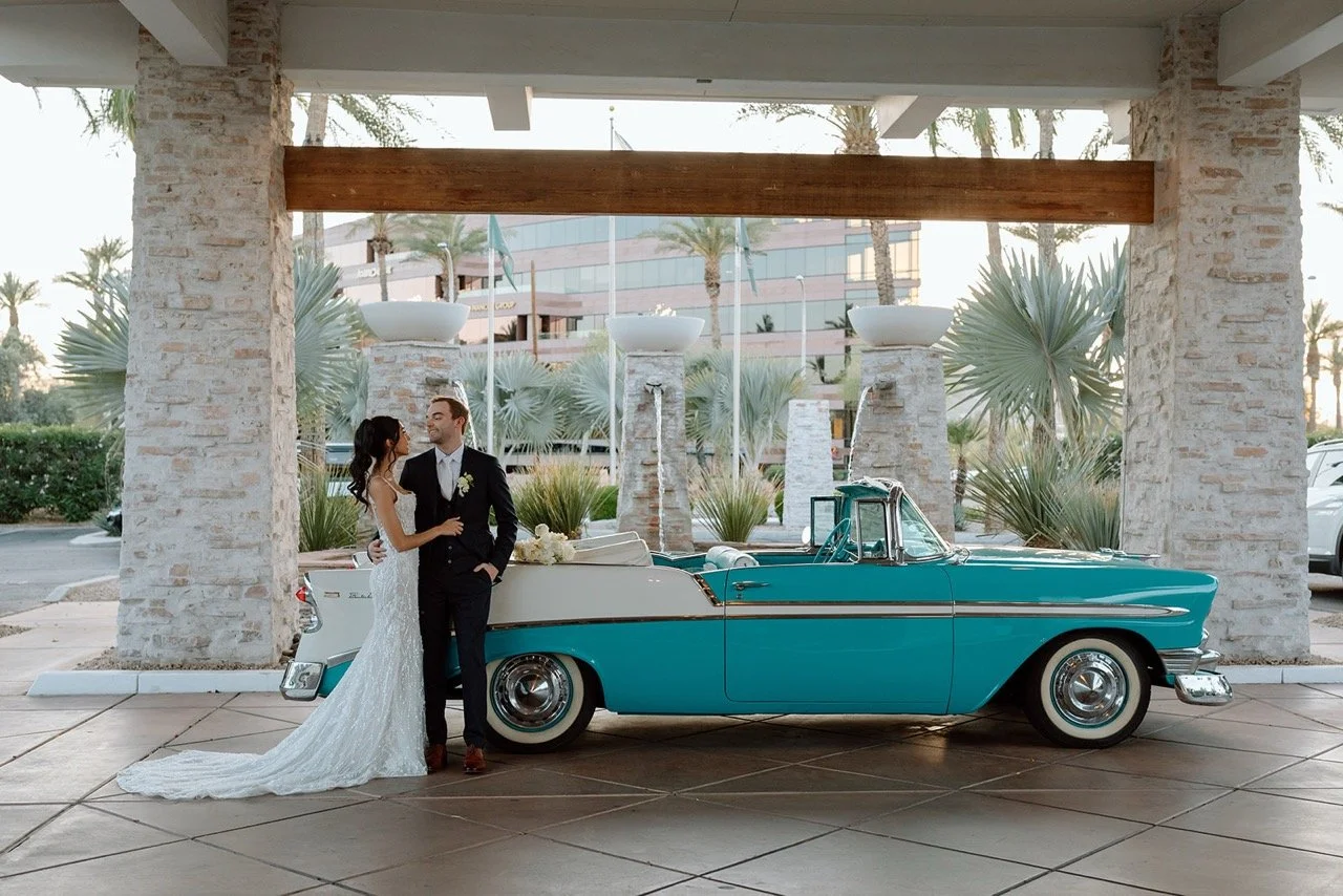 A bride and groom standing beside a vintage turquoise convertible car at a wedding venue with stone pillars and desert plants.