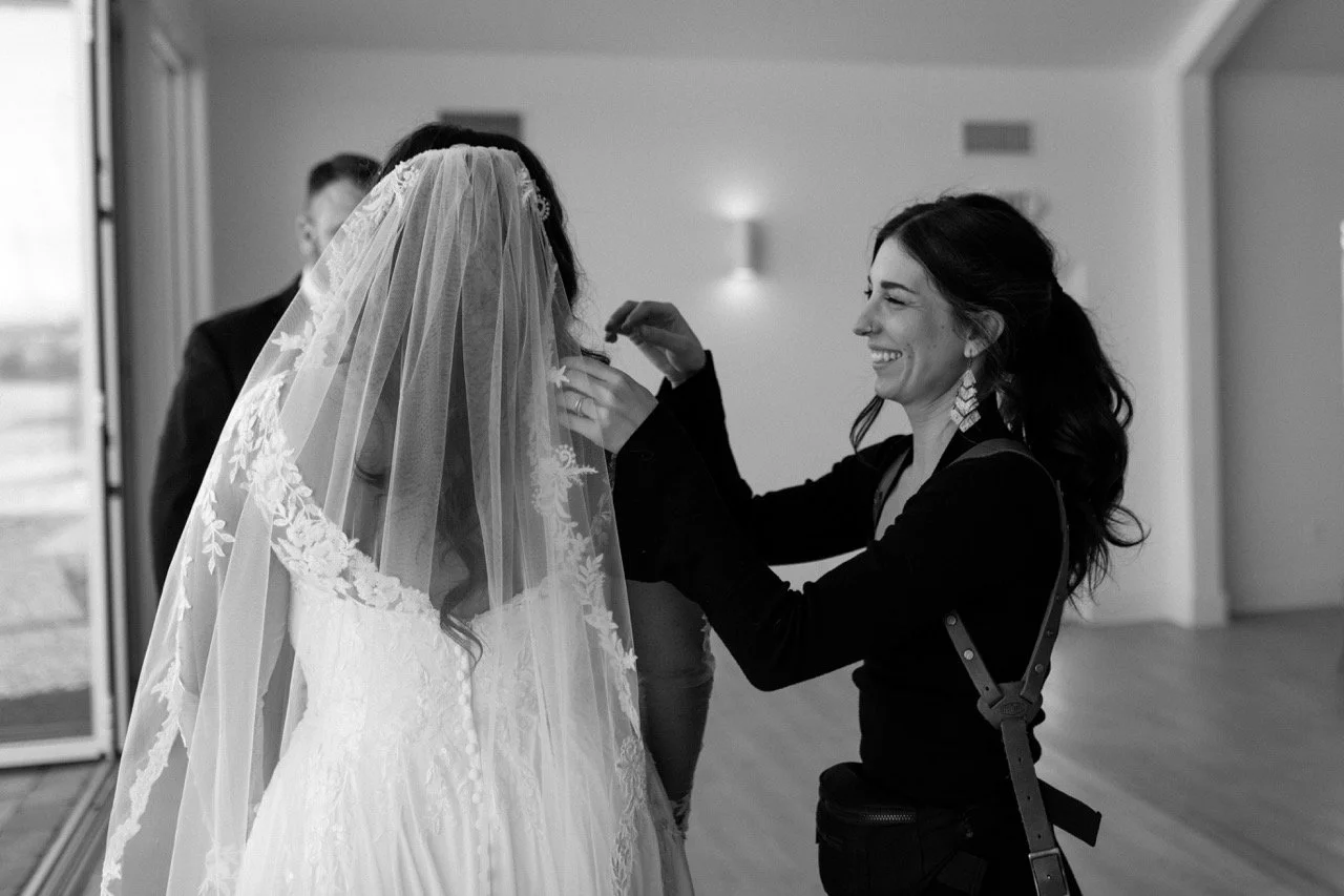 A bride wearing a wedding gown and veil is having earrings put on by a smiling woman, likely a stylist or friend, in a bright room with a large window.