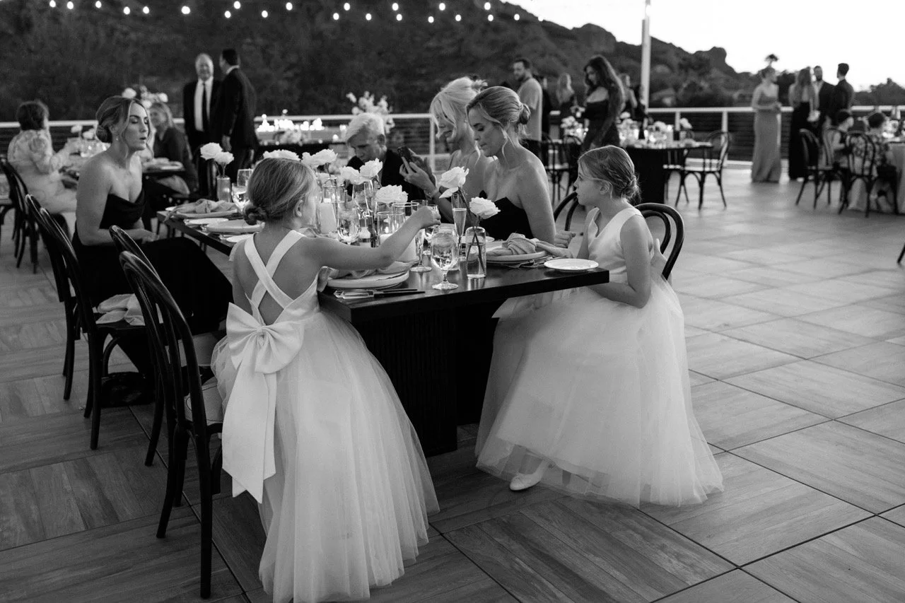 Guests, including children in fancy dresses, dining at an outdoor wedding reception table with fine tableware and floral centerpieces.