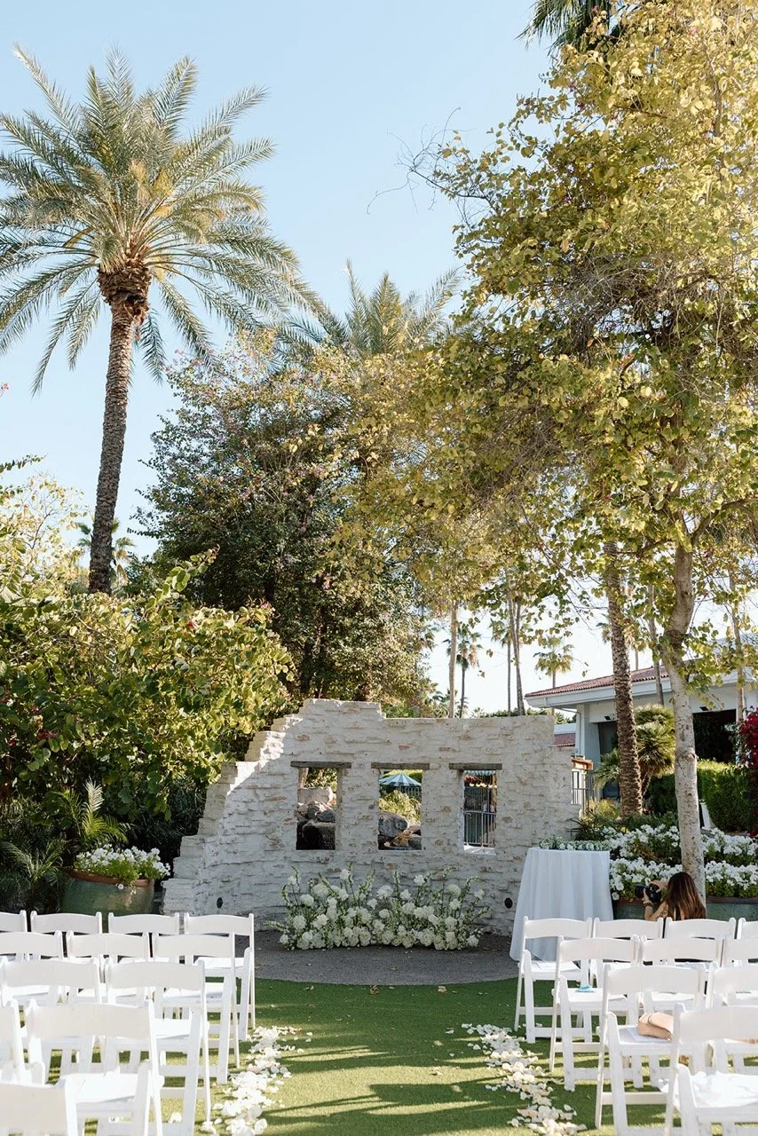 Outdoor wedding ceremony setup with white chairs, floral decor, and palm trees under a clear sky.