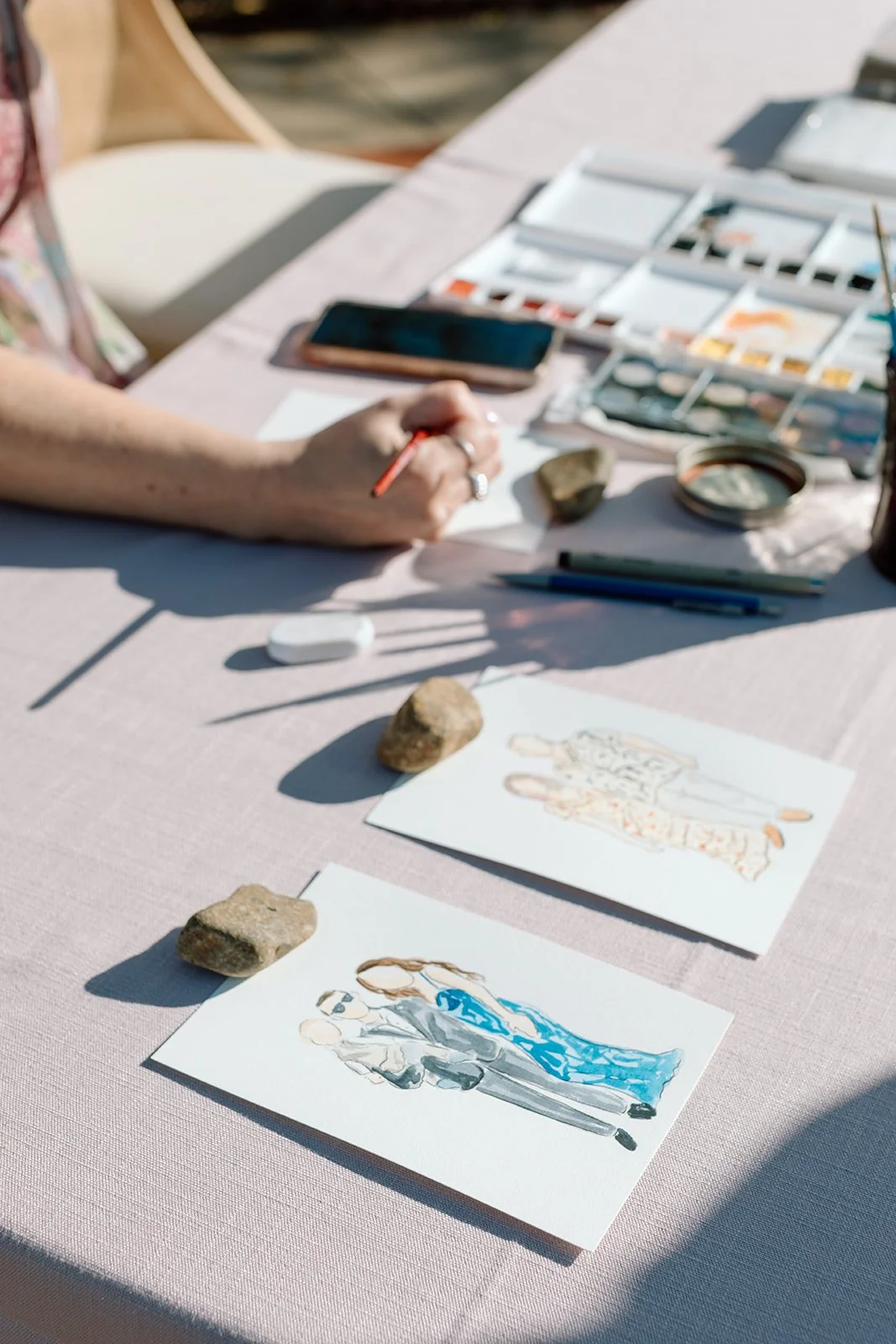 Artist creating watercolor fashion illustrations at an outdoor table with paint palette, brushes, smartphone, and rocks for decoration.