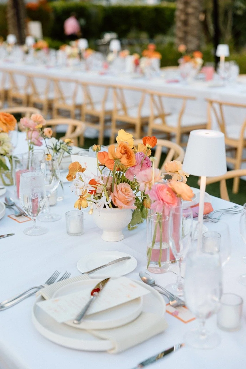 Elegant outdoor event table decorated with pink and peach flowers, white tablecloths, and place settings including plates, silverware, wine glasses, and small candles.