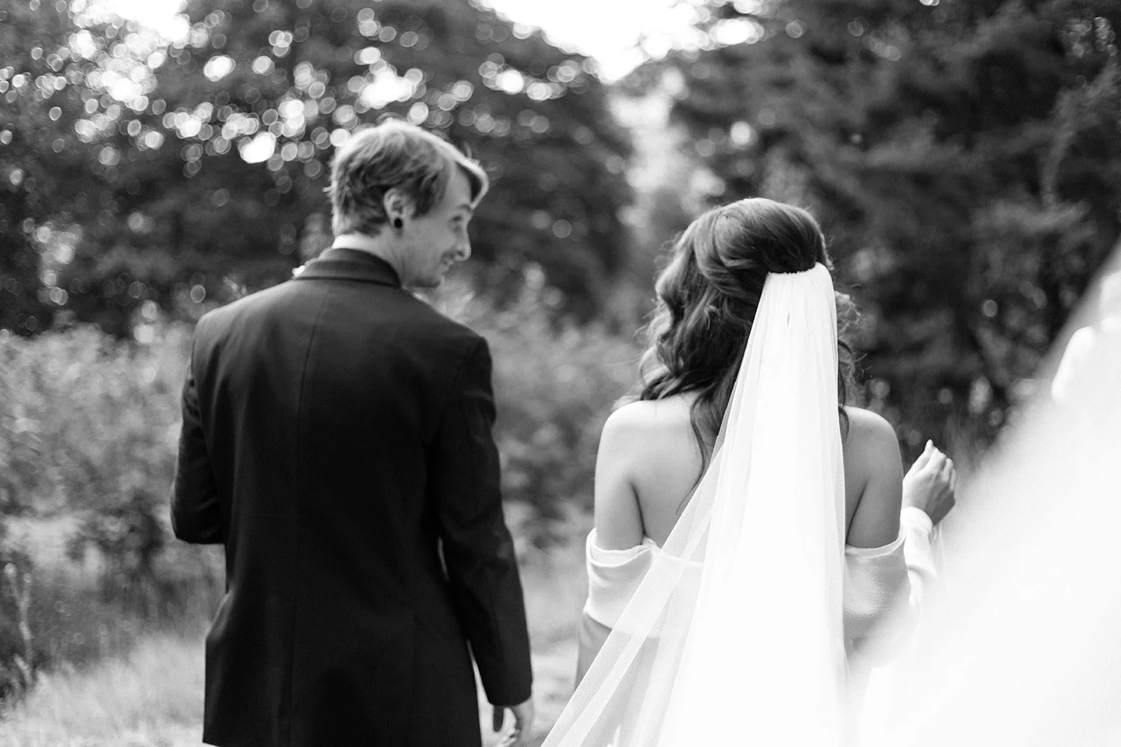 A black and white photograph of a bride and groom standing outdoors, with the groom looking at the bride. The bride has long hair with a veil, and the groom is wearing a suit. They are surrounded by trees, and the scene appears to be part of a weddin