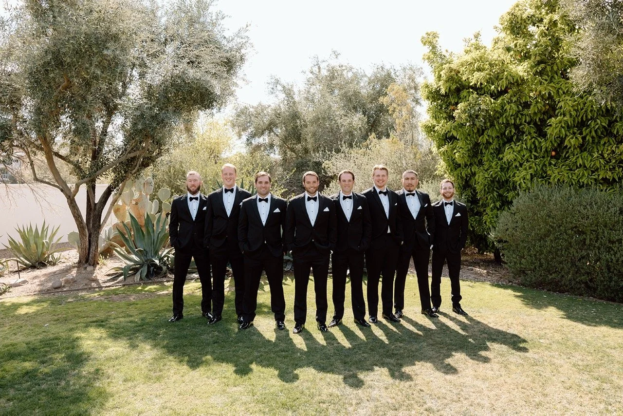 A group of nine men in tuxedos standing outdoors on grass, smiling, with trees and bushes in the background.