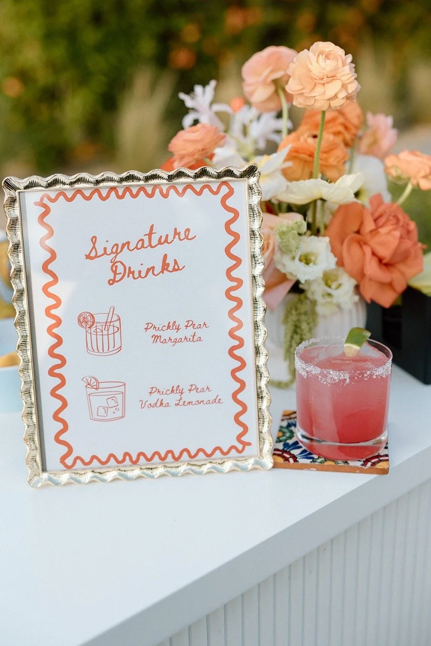 A pink cocktail with a lime wedge garnish on a table with a decorative sign that reads 'Signature Drinks,' listing a 'Prickly Pear Margarita' and a 'Prickly Pear Vodka Lemonade,' with a background of peach and white flowers.