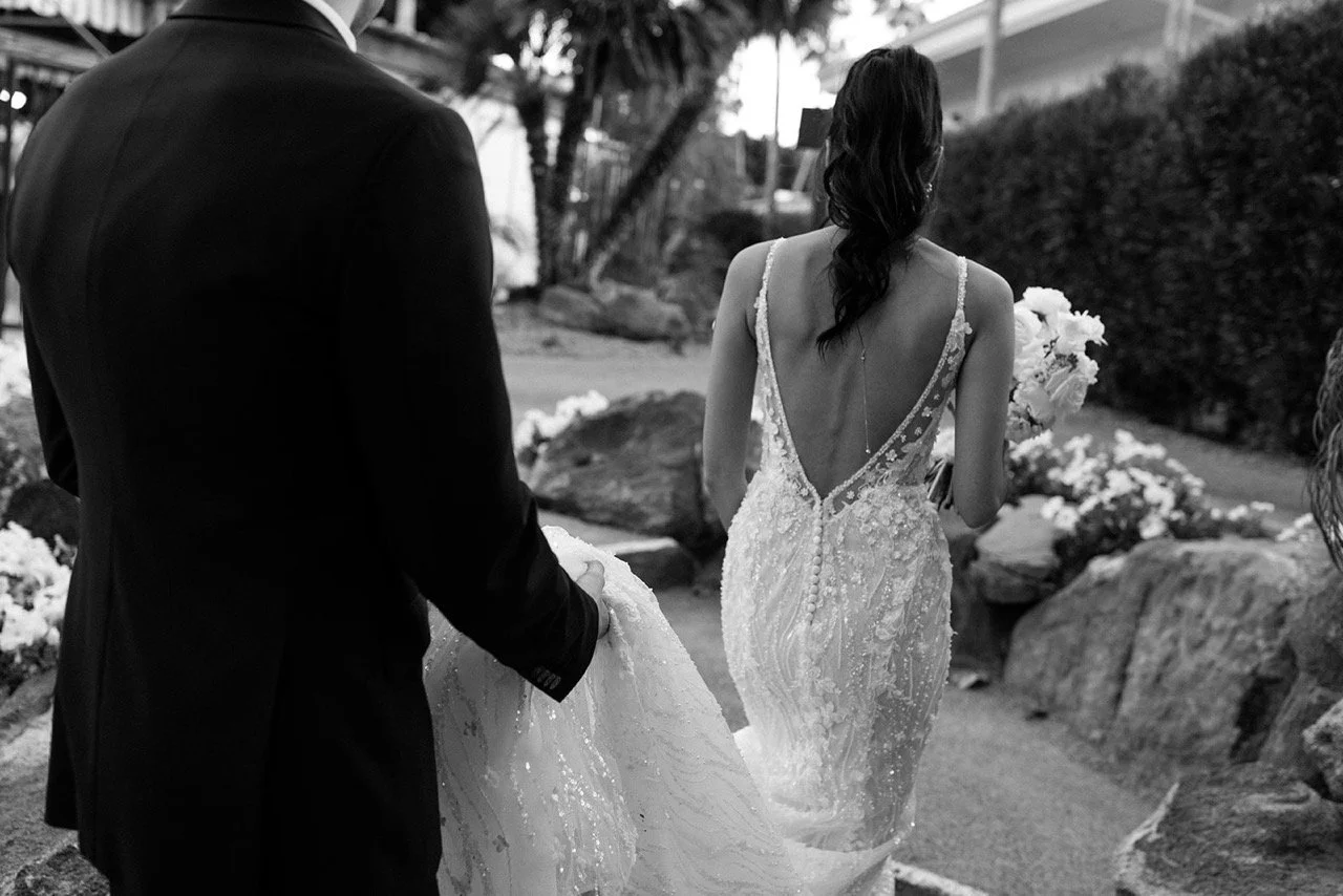 Black and white photo of a bride in a wedding dress walking outdoors, holding a bouquet, with a man in a dark suit assisting with her gown.