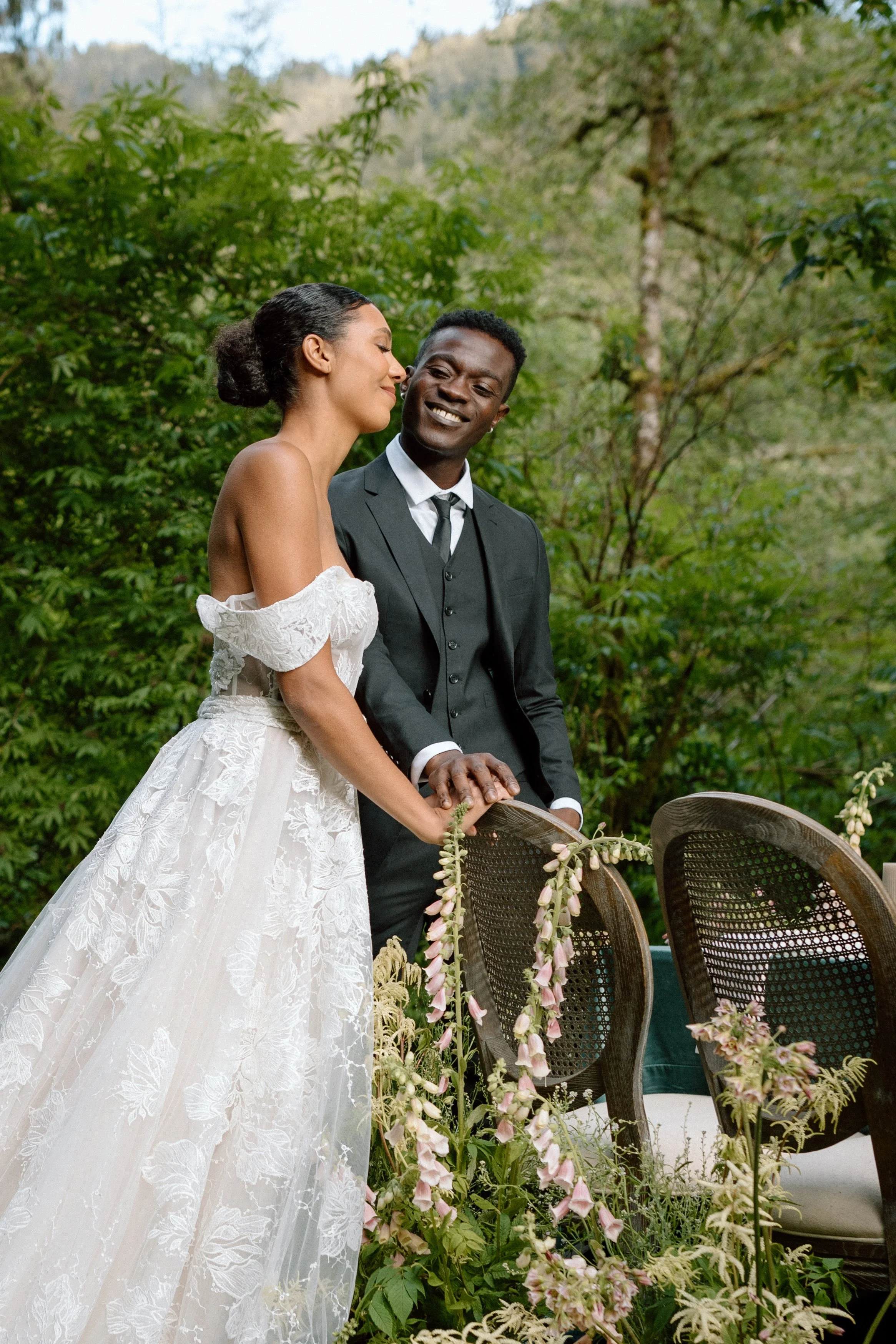 A bride and groom standing outdoors, holding hands, smiling, with greenery and trees in the background.