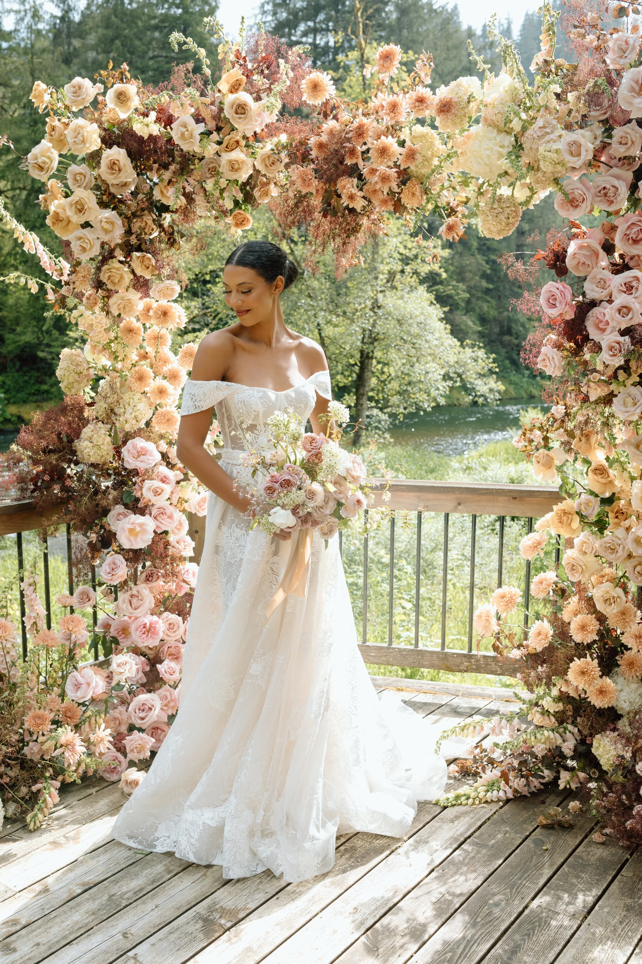 A bride in a white wedding dress standing under a floral arch made of pink and peach roses and other flowers on a wooden deck overlooking a river and green trees.