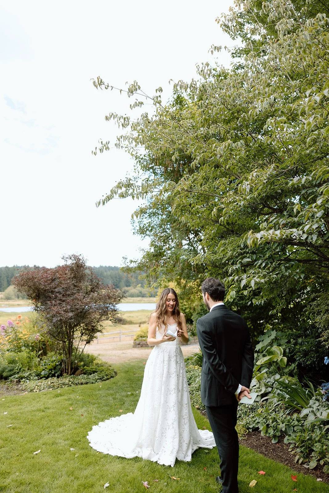 A bride in a white wedding dress is smiling and holding a piece of paper, standing in a garden with greenery and trees. A groom dressed in a black suit stands nearby with his back to the camera, holding a paper behind his back. The scene appears to b