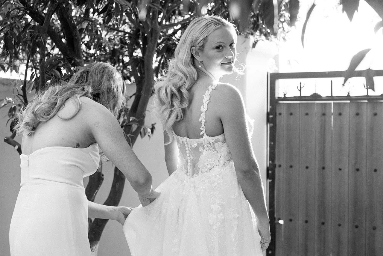 A bride in a floral lace wedding dress turning to smile at the camera while another woman adjusts her dress, outdoors with a tree and a wooden gate in the background.