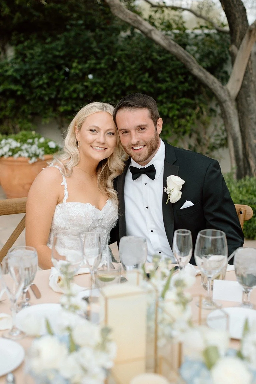 A newlywed couple sitting at a table outdoors, smiling at the camera, bride in a white lace wedding dress and groom in a black tuxedo with a bowtie and white boutonniere.