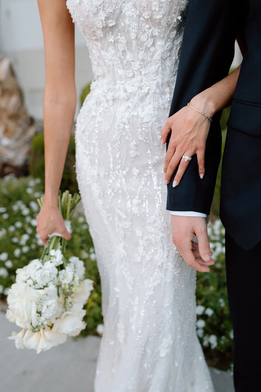 Close-up of a bride and groom holding hands during a wedding, with the bride wearing a white, intricately beaded wedding gown and holding a bouquet of white flowers, and the groom dressed in a black suit