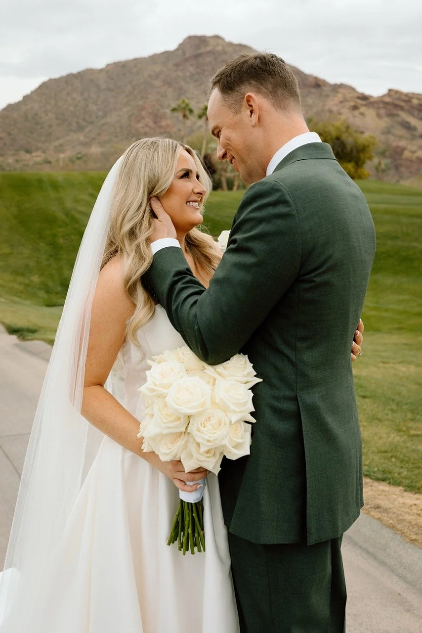A bride and groom smiling and gazing at each other outdoors on a wedding day, with the bride holding a bouquet of white roses