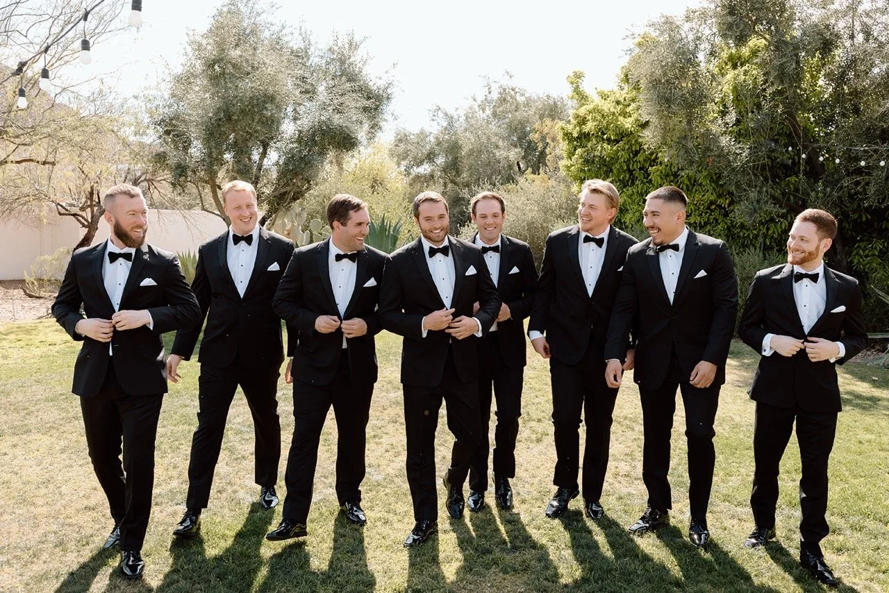 Group of nine men in black tuxedos with bow ties standing outdoors on a grassy area, smiling and talking to each other.