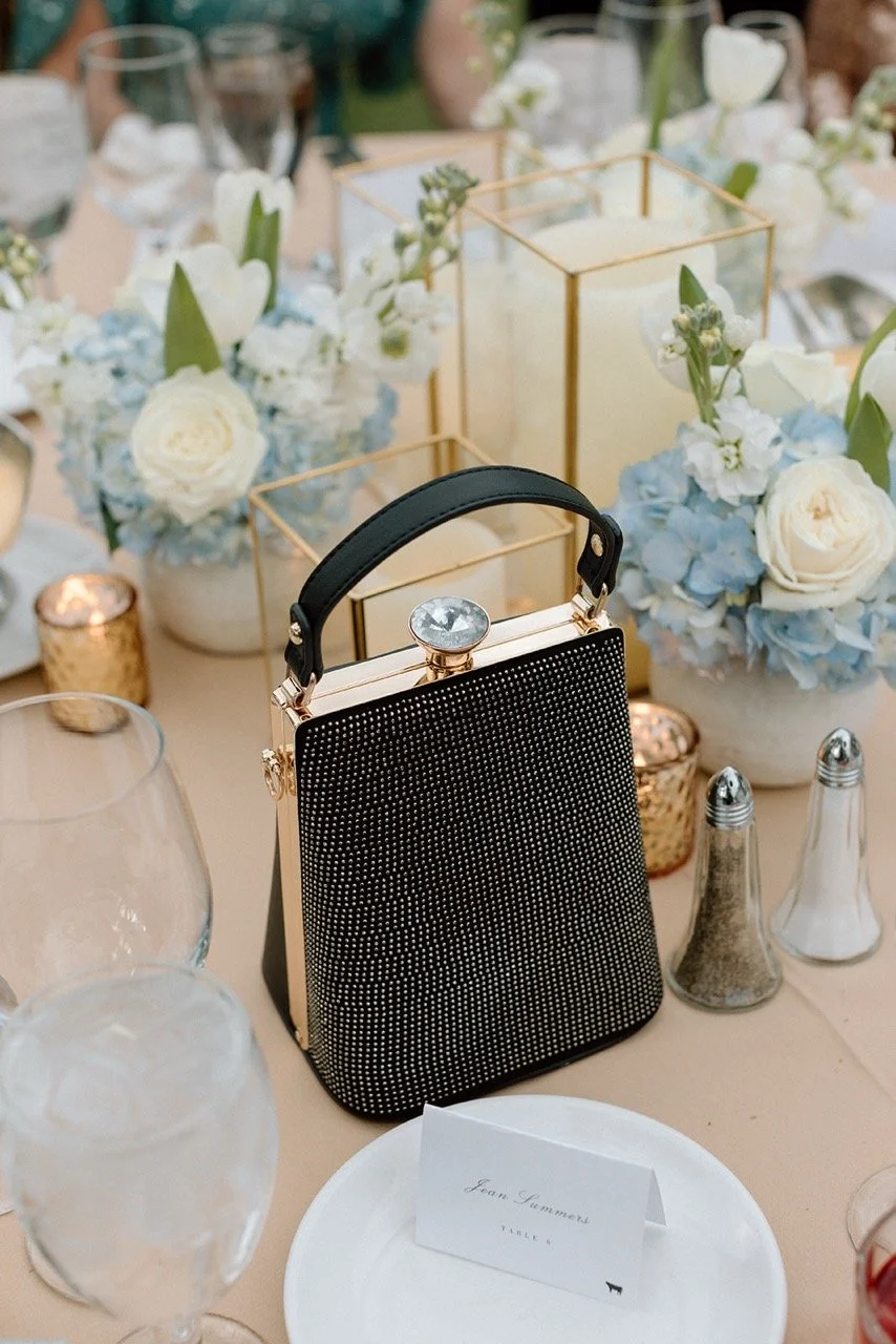 A wedding or formal event table setting with a black handbag decorated with silver rhinestones, surrounded by white and blue floral centerpieces, glassware, candles, and a place card for Joan Sumner.