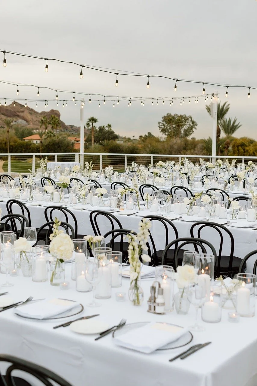 Outdoor wedding reception setup with long white tables, black chairs, and string lights, featuring white floral centerpieces and candles.
