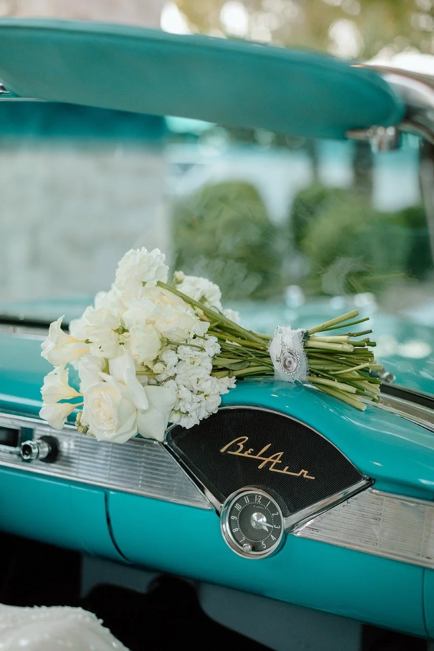 A vintage light blue car dashboard with a bouquet of white flowers lying on it and a small clock. The dashboard has a black and gold nameplate with the word 'Bel-Aire'.
