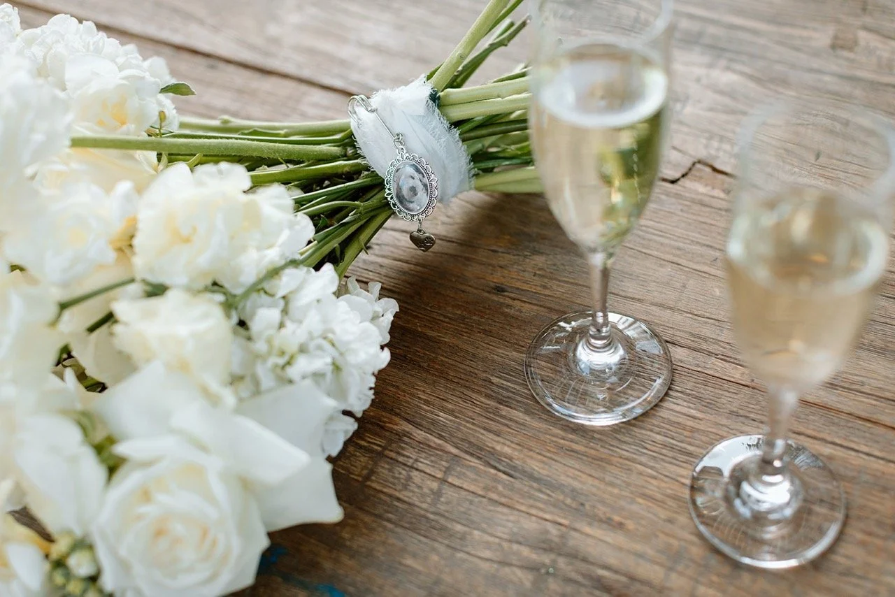 A bouquet of white flowers with a silver pendant necklace on a wooden table, accompanied by three glasses of champagne or sparkling wine.