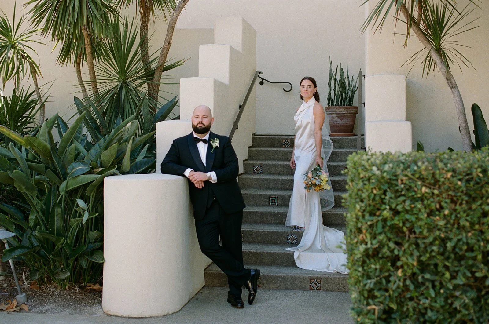 A man in a black tuxedo and a woman in a white dress pose on outdoor steps with tropical plants and white walls.