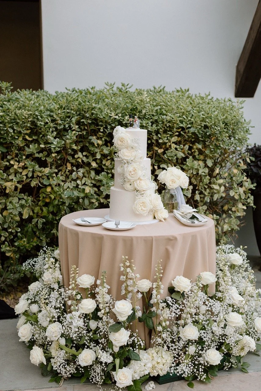 Wedding cake with white roses and floral decorations on a round table, surrounded by white flowers, outdoors.